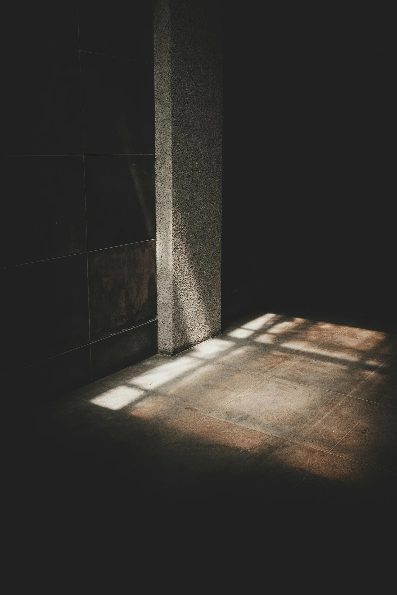 Lumière du soleil qui passe à travers une fenêtre, créant des motifs de lumière sur le sol et un mur dans une pièce sombre.